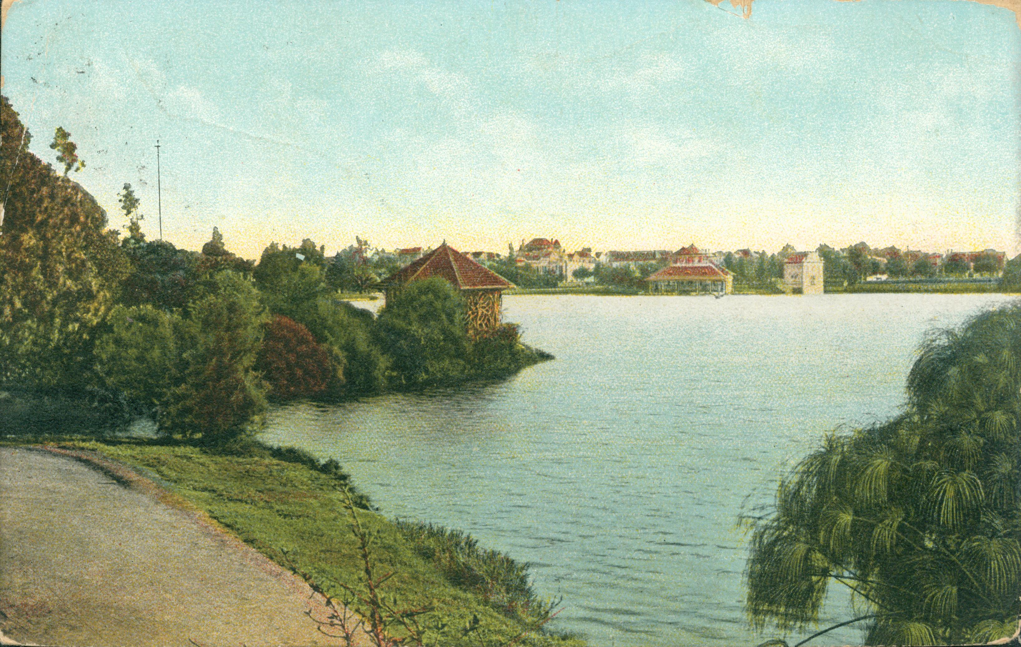 This postcard shows a path beside a lake with a gazebo on the left.